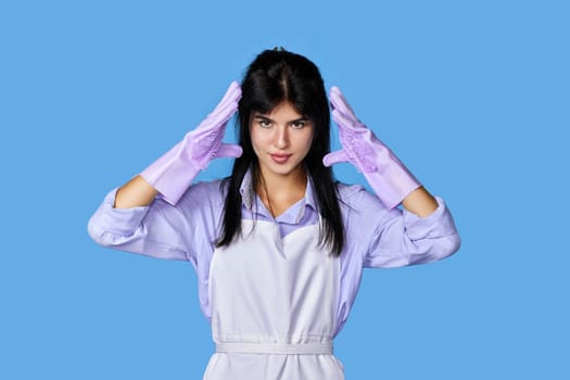 upset woman in rubber gloves and cleaner apron with stress headache looking at camera isolated on blue background. cleaning