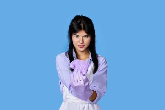 beautiful caucasian woman in violet rubber gloves and cleaner apron ready to clean on blue background.