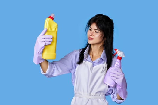 beautiful woman in violet rubber gloves and cleaner apron chooses between two cleaning products on blue background.