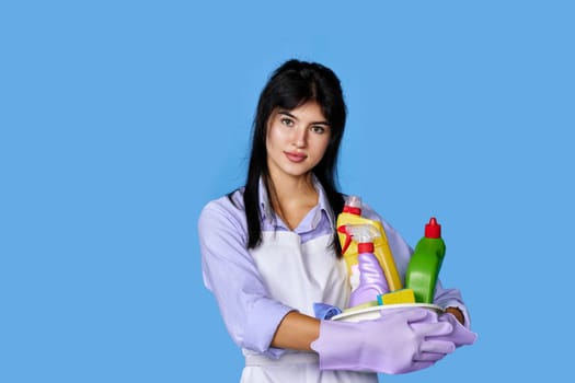 smiling caucasian woman in gloves holding bucket with cleaning supplies isolated on blue background.