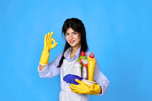 happy caucasian woman in gloves and cleaner apron holding bucket of detergents and showing Ok gesture isolated on blue background.
