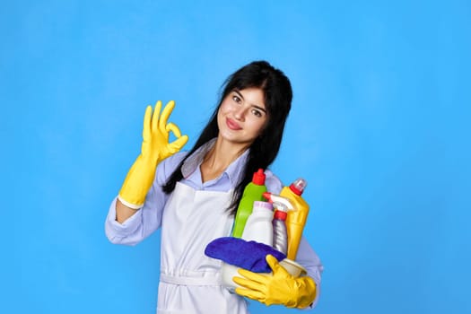 pretty woman in yellow rubber gloves and cleaner apron holding bucket of detergents and showing Ok gesture isolated on blue background.