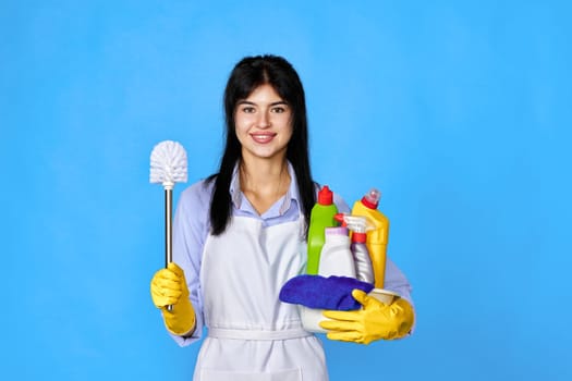 pretty woman in yellow rubber gloves and cleaner apron holding bucket of detergents and toilet brush on blue background.