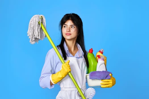 pretty woman in yellow rubber gloves and cleaner apron holding bucket of detergents and mop on blue background.