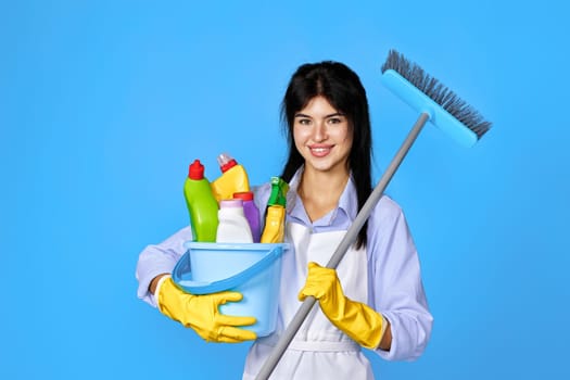 young smiling woman in yellow rubber gloves and cleaner apron holding bucket with cleaning supplies and broom on blue background.