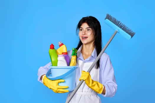 surprised pretty woman in yellow rubber gloves and cleaner apron holding bucket of detergents and broom on blue background.