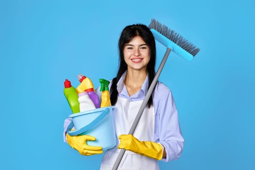 happy woman in gloves and cleaner apron holding bucket of detergents and broom on blue background.