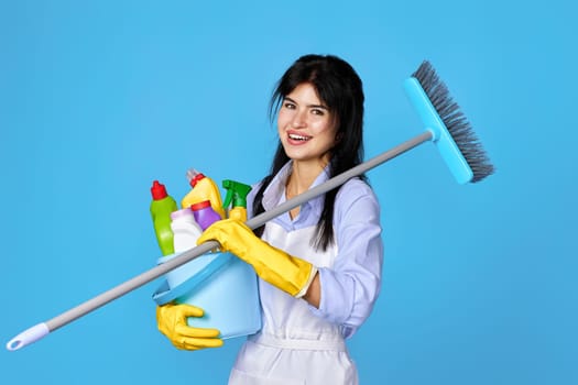 cheerful woman in yellow rubber gloves and cleaner apron holding bucket of detergents and broom on blue background.