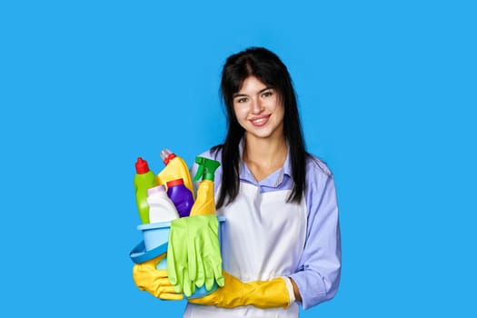 smiling caucasian woman in gloves holding bucket with cleaning supplies isolated on blue background.