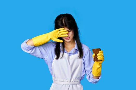 tired woman gloves and cleaner apron with sponge on blue background