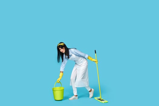 woman in rubber gloves and cleaner apron washing floor with mop on blue background. Full length
