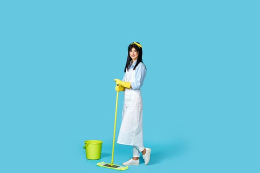 woman in rubber gloves and cleaner apron washing floor with mop on blue background. Full length