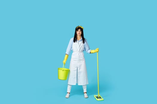positive cheerful professional cleaner woman in rubber gloves and cleaner apron washing floor with mop on blue background. Full length