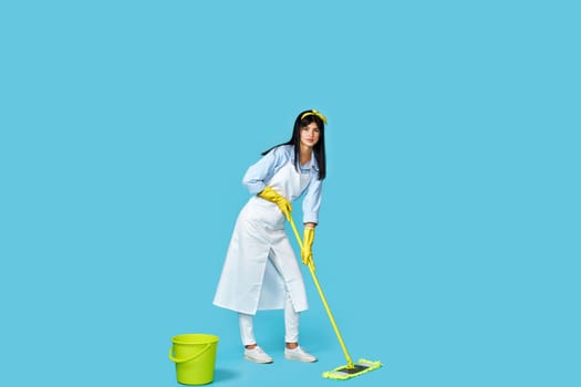 woman in rubber gloves and cleaner apron washing floor with mop on blue background. Full length