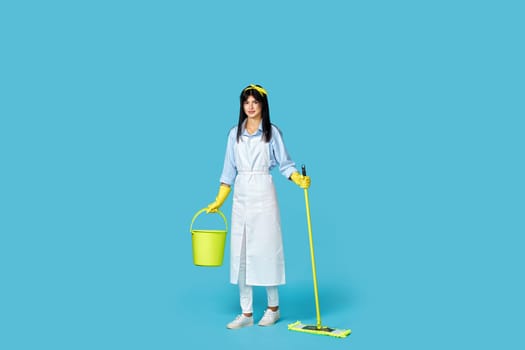 professional cleaner woman in rubber gloves and cleaner apron holding mop and bucket on blue background. Full length