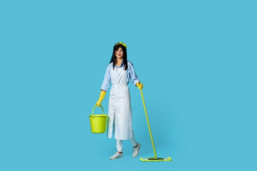 professional woman in rubber gloves and cleaner apron holding mop and bucket and feeling happy and carefree on blue background. Full length