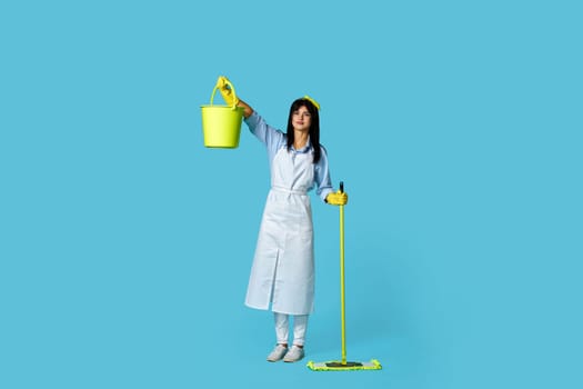 charming woman in rubber gloves and cleaner apron holding mop and bucket on blue background. Full length