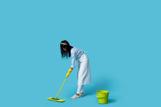 woman in rubber gloves and cleaner apron washing floor with mop on blue background. Full length