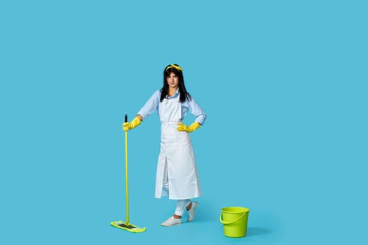 professional cleaner girl in rubber gloves and cleaner apron washing floor with mop on blue background. Full length