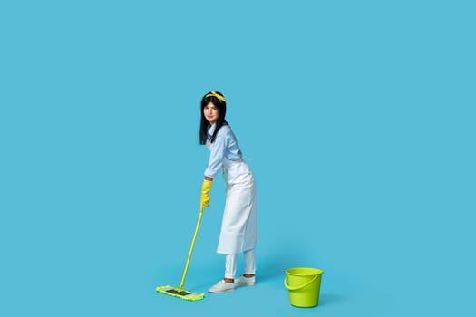 cheerful woman in rubber gloves and cleaner apron washing floor with mop on blue background. Full length