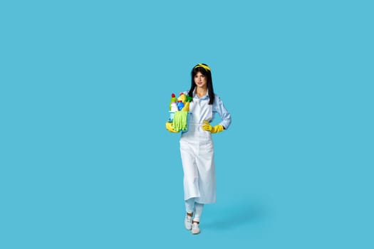 happy cheerful woman in rubber gloves and cleaner apron with cleaning supplies on blue background. Full length