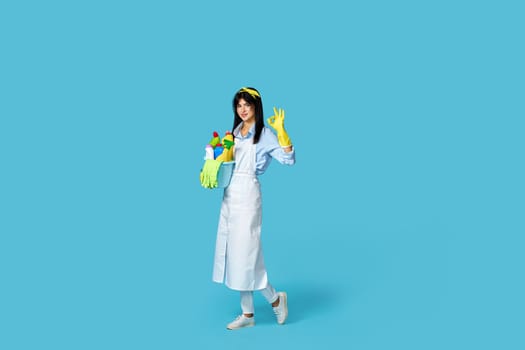 pretty cheerful woman in rubber gloves and cleaner apron with cleaning supplies showing ok gesture on blue background. Full length