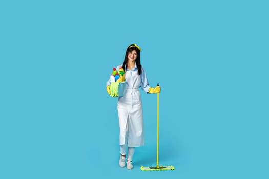 professional cleaner woman in rubber gloves and cleaner apron with cleaning supplies and mop on blue background