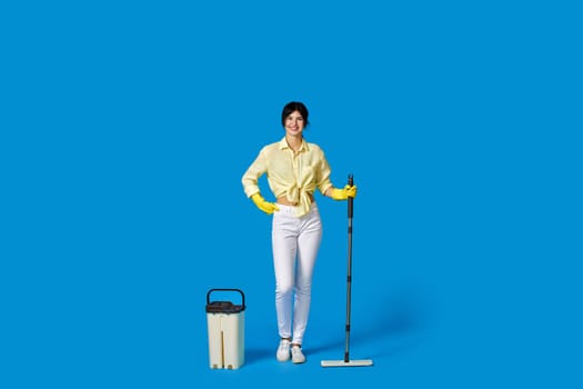 cheerful housewife woman in yellow rubber gloves washing floor with mop on blue background. Full length