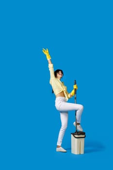funny happy housewife girl in yellow rubber gloves washing floor with mop and dancing on blue background. Full length
