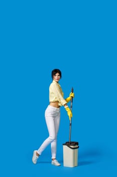 beautiful housewife girl in rubber gloves washing floor with mop on blue background. Full length