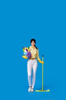 concenteated serious girl in rubber gloves housekeeping and cleaning with mop and holding bucket of detergents on blue background. Full length