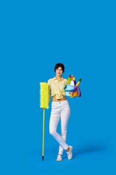 concenteated serious housewife girl in rubber gloves housekeeping and cleaning with mop and holding bucket of detergents on blue background. Full length