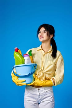 caucasian brunette woman in yellow gloves holding bucket with cleaning supplies isolated on blue background.