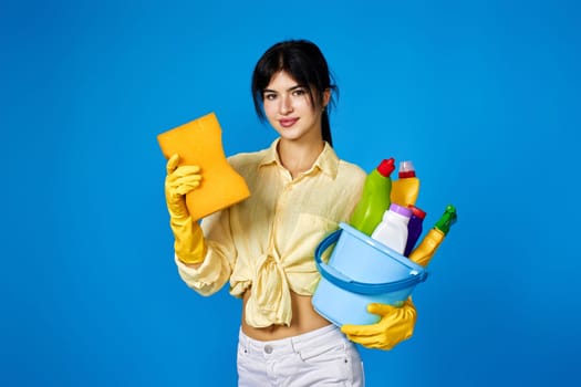 caucasian happy housewife in yellow gloves holding sponge and bucket with cleaning supplies isolated on blue background.