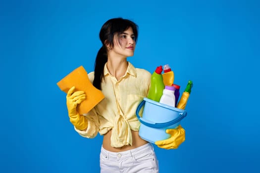 caucasian brunette woman in yellow gloves housekeeping and cleaning with sponge and bucket with cleaning supplies isolated on blue background.