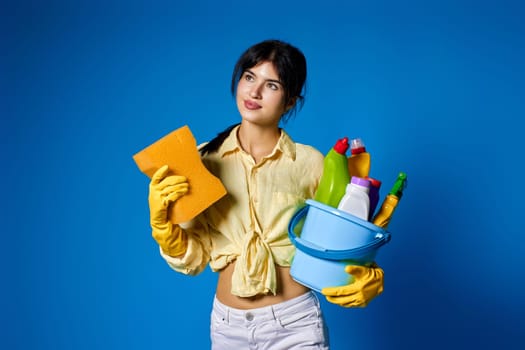 caucasian happy housewife in yellow gloves holding sponge and bucket with cleaning supplies isolated on blue background.