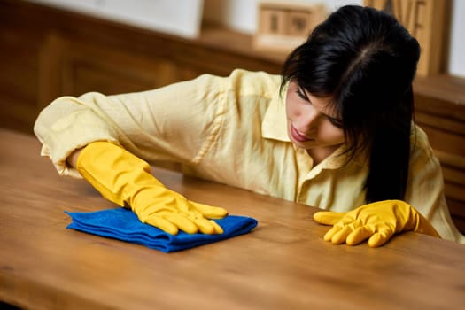 brunette woman in yellow gloves using a rag while cleaning furniture at home