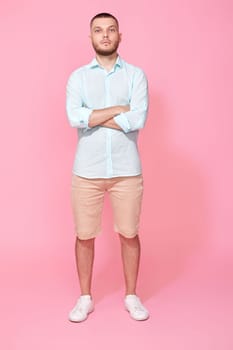 young caucasian man wearing blue t-shirt holding hands crossed looking camera isolated on pink background. Full length