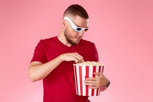 Young caucasian man in 3d glasses eating popcorn on pink background