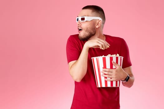 Young caucasian man in 3d glasses eating popcorn on pink background