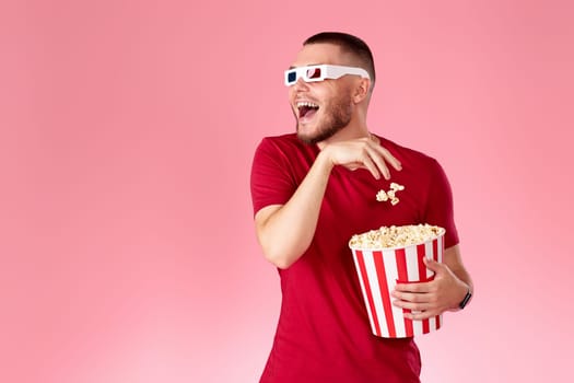 Young caucasian man in 3d glasses eating popcorn on pink background