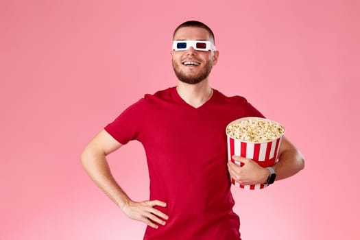 Young caucasian man in 3d glasses eating popcorn on pink background