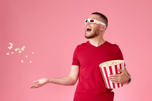 young handsome man in 3d glasses and t-shirt holding popcorn box and throwing up popcorn on pink background