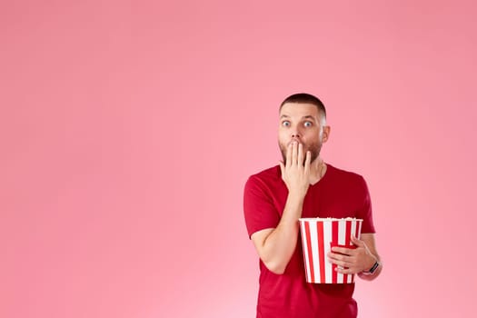 Shocked scared young caucasian man eating popcorn on pink background. copy space