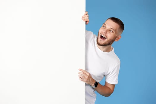 Portrait of friendly man in casual t-shirt peeking out from behind advertisement whiteboard with empty copy space on blue background