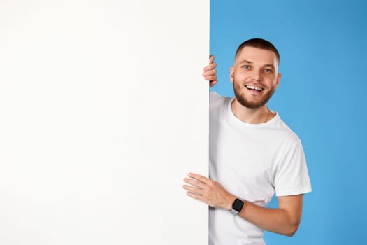 Portrait of cheerful bearded man standing by white empty advertising board isolated on blue background
