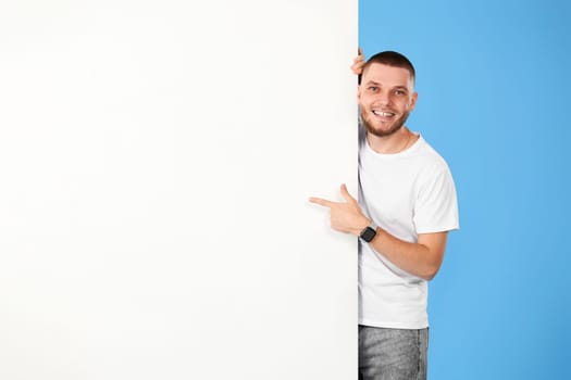 Portrait of cheerful bearded man pointing at blank advertising board with empty copy space isolated on blue background. mock-up