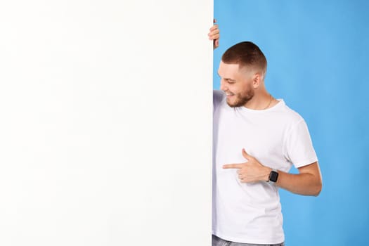 Portrait of friendly man point finger empty space banner isolated on blue background. mock-up
