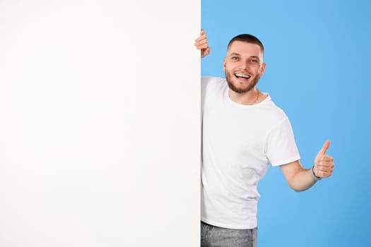 Portrait of cheerful bearded man peeking out from behind advertisement whiteboard and showing thumb up isolated on blue background