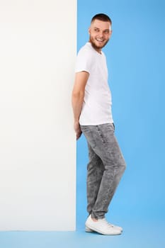 Portrait of friendly guy in casual t-shirt leaning on empty white placard for advertisement on blue background